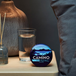 A lifestyle image of a Camino Midnight Blueberry Tin sitting on a night table next to a bed.