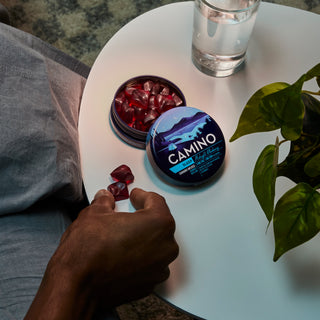 A lifestyle image from a mans perspective of a night table next to a bed. The man is reaching his hand to a Camino Midnight Blueberry Gummy sitting on the night table next to a slightly opened Camino Midnight Blueberry Tin.