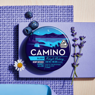 A flatlay photograph of the Camino Midnight Blueberry tin with sprigs of lavender, chamomile flowers and blueberries around the tin. 
